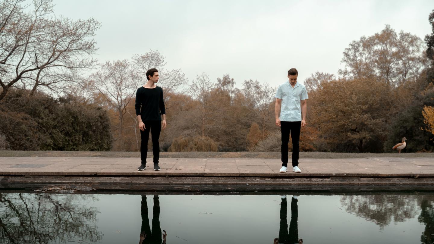 Two brother stand at the edge of a pond. One stares at his reflection. The other stares across at his brother.