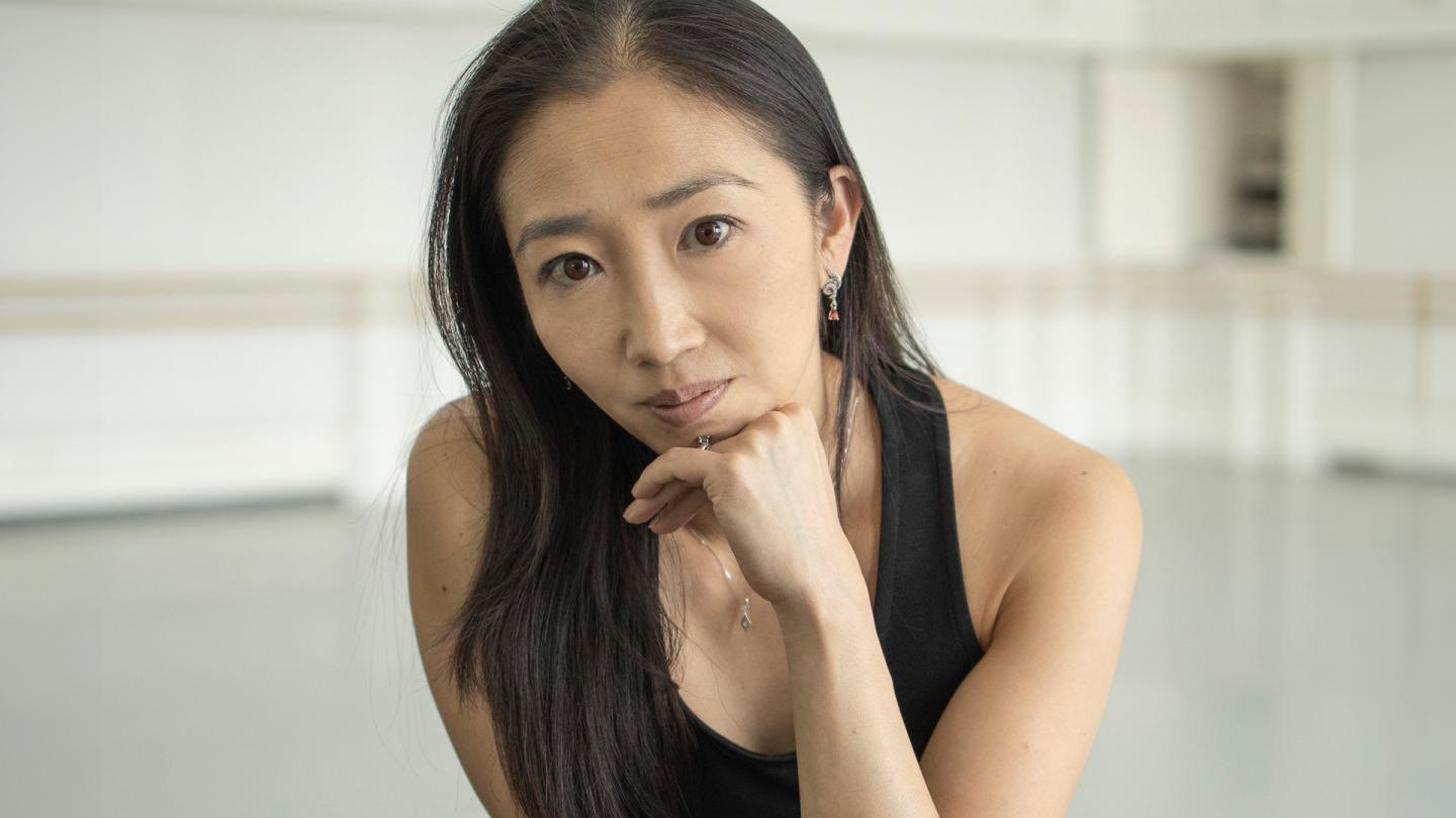 Woman with brown eyes, long dark hair, wearing a black vest resting their chin on her hand as she leans forward looking directly at the camera