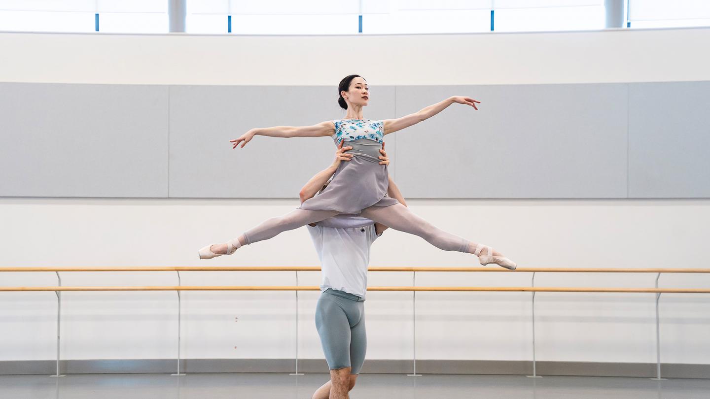 One dancer lifts another by the waist above his head, she holds her arms and legs stretched away from her body