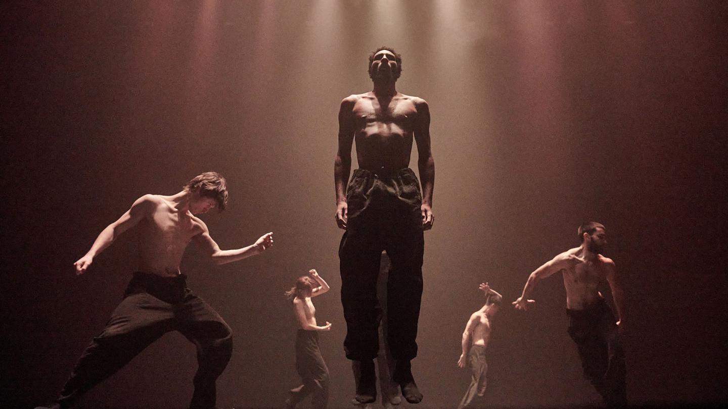 Five dancers in hazed light striking firm poses, one central and lower half of his face cast in shadow