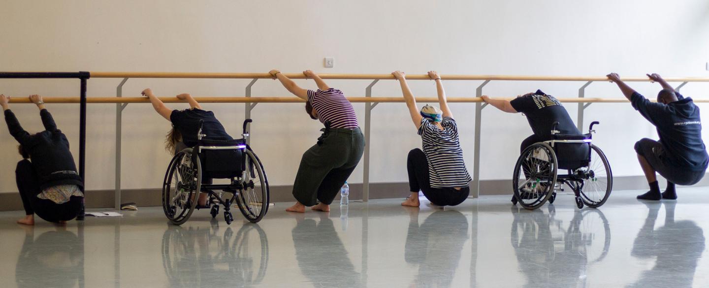 Two people in wheelchairs and three people standing, all stretching against a ballet barre