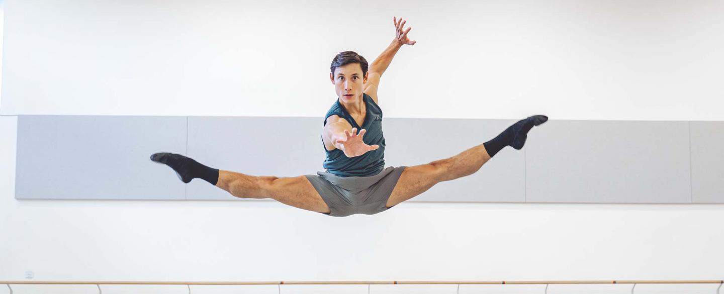 A male dancer jump with both legs extended in a mid-air split while reaching for the camara