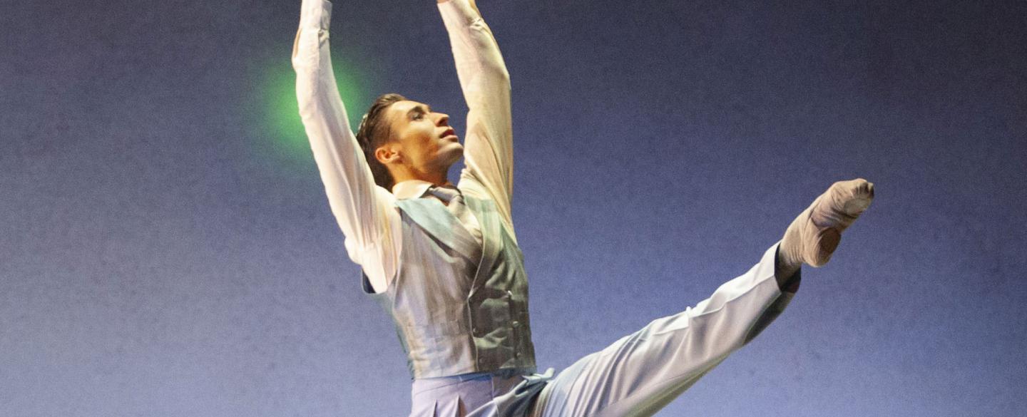 Male dancer stood against a blue stage with a green light behind him. His arms are raised and one leg is extended.