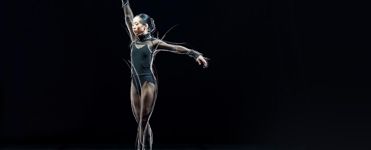 Dancer in a black leotard stands en pointe against a black background with one arm extended above her and one to her side.