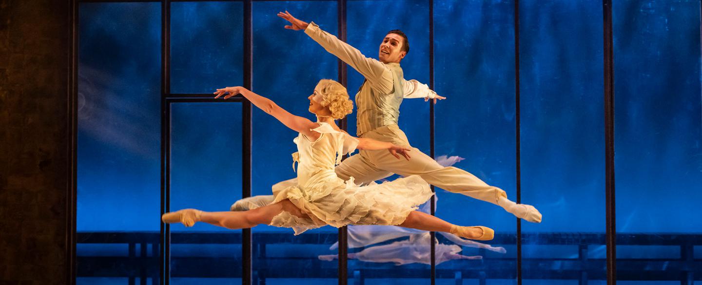 A male and female dancer both performing grand jetés joyously while wearing while clothes against a blue background