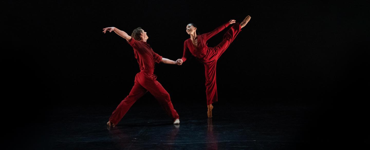 Male and female dancer in red jump suits standing on point with one leg raised high.