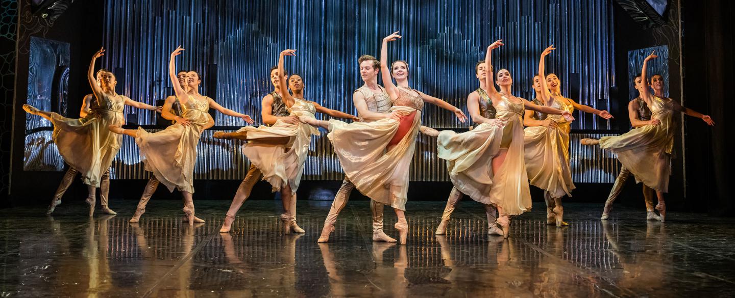 Dancers, dressed in pale gold costume, paired with the male holding the female by the waist for stability as she in en pointe ion one foot and in arabesque.