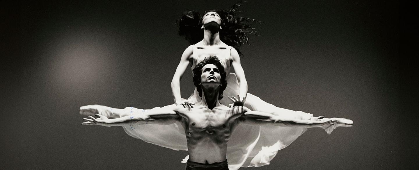 Male dancer, shirtless, standing with his arms wide and a female dancer in a white dress is behind him, holding herself up on his shoulders with her legs split in parallel with the male dancers arms