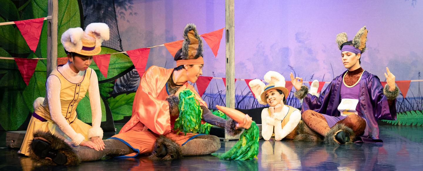 Dancers dressed as bunnies. One dancer checking another dancers leg, middle dancer holding two carrot stick pom poms, another dancer gazing in adoration and the last dancer meditating