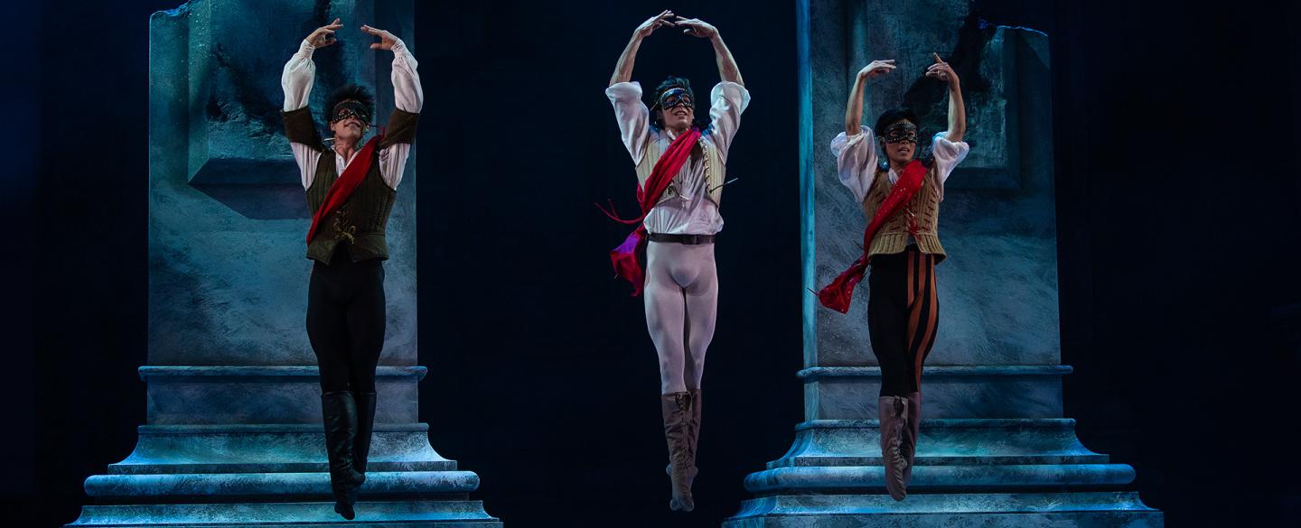 Three dancers in masks wearing a red sash jump with their feet together and their arms raised above their head