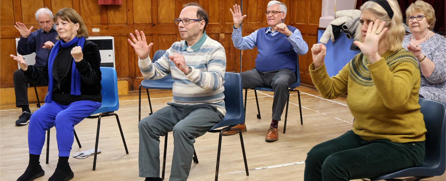 A group of people sat in plastic chairs raising their hands copying an unseen instructor