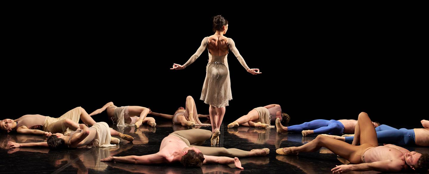 A group of dancers lie on the ground, apparently unconscious, while another stands and walks through the fallen bodies