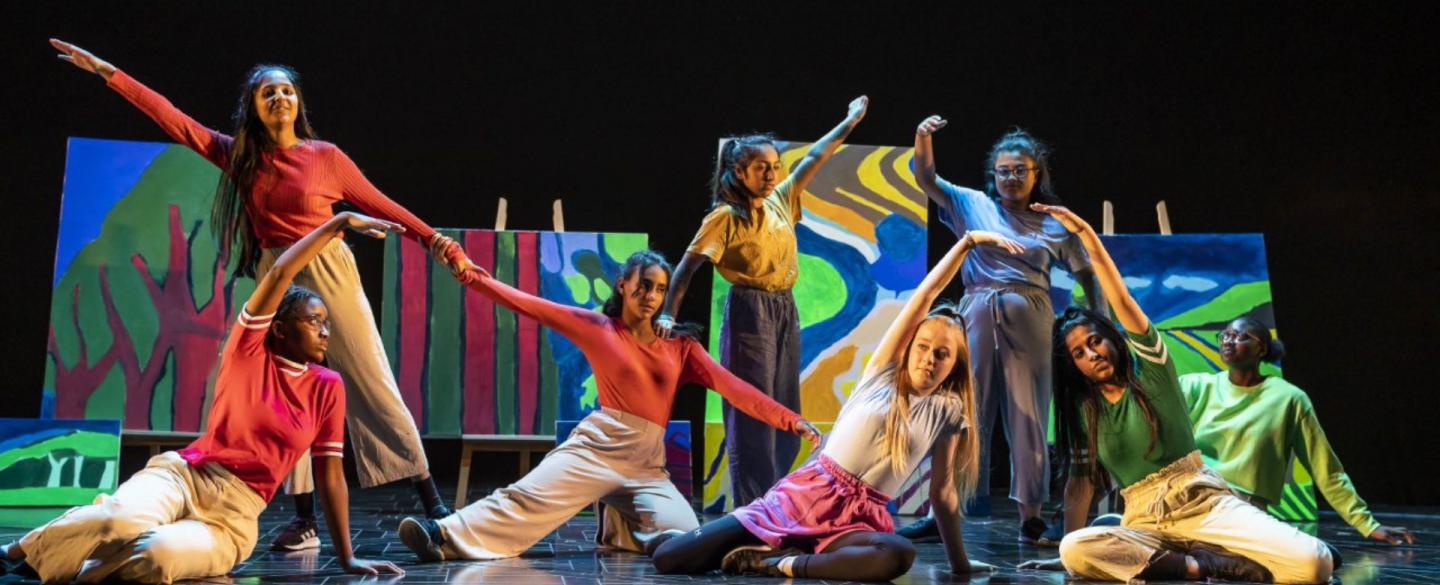 Performers wearing different colours pose on a colourful stage. Some are sat and some are standing and all reach their arm outwards in different direction.