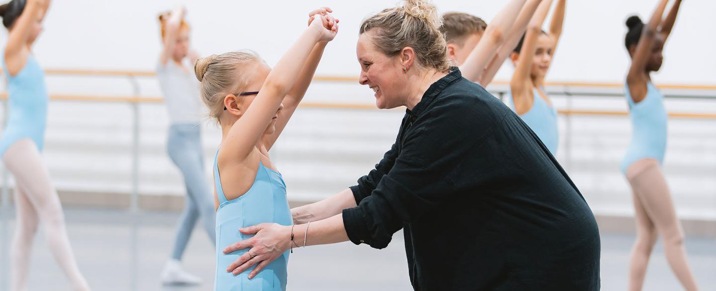 CAT Student in uniform pale blue leotard holds her hands above her head while her teacher helps her posture and smiles with encouragement