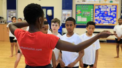 A group of pupils in a school hall watch as a dance teacher addresses them. The dancer teacher's back is to the camera, she is pointing across to the right of the image.