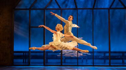 A male and female dancer both performing grand jetés joyously while wearing while clothes against a blue background