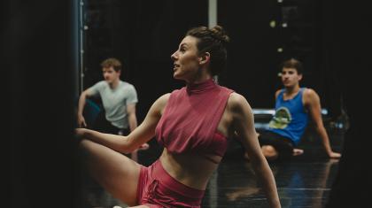 Choreographer wearing pink, sat on the floor leaning back on one arm, while talking with someone out of frame