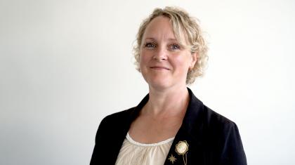 Woman with short blonde hair, wearing a white top and black jacket, smiles at the camera