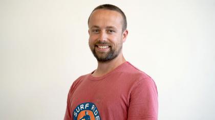 MAn smiles at the camera, he has short dark hair, a beard, and is wearing a pink T-shirt with the text Surf Riders printed on it.