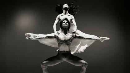 Male dancer, shirtless, standing with his arms wide and a female dancer in a white dress is behind him, holding herself up on his shoulders with her legs split in parallel with the male dancers arms