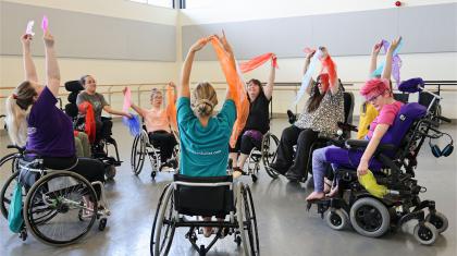 Group of dancers in wheelchairs rehearse in studio reach high holding colourful ribbons and fabric.