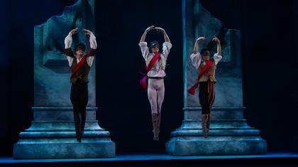 Three dancers in masks wearing a red sash jump with their feet together and their arms raised above their head