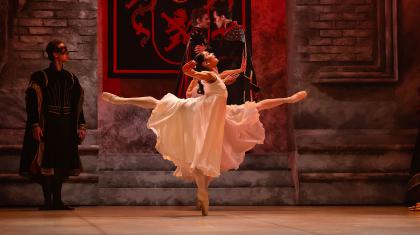 Dancer dressed in a white flowing dress dances elegantly, one leg raised behind her perpendicular to the floor while en pointe with the other