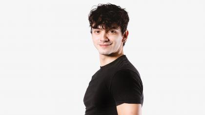 Man with short dark hair, wearing a black T-shirt, stand side-on to the camera but looks directly at it, smiling