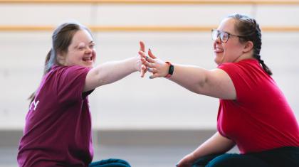 Two dancers in an Ability class sit across from each other on the floor, touching hands and smiling