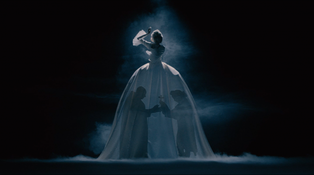 A woman in a huge white dress stands in the dark as the shadows of two men are projected onto her dress.