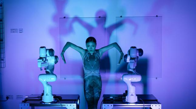 A dancer stood between two robots in purple lighting